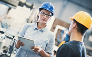 Facility management, technical facility management, woman wearing work clothes and a hardhat pointing at a computer screen, slightly blurred mand wearing wok clothes holding a pen and pad, blurred factory in the background 